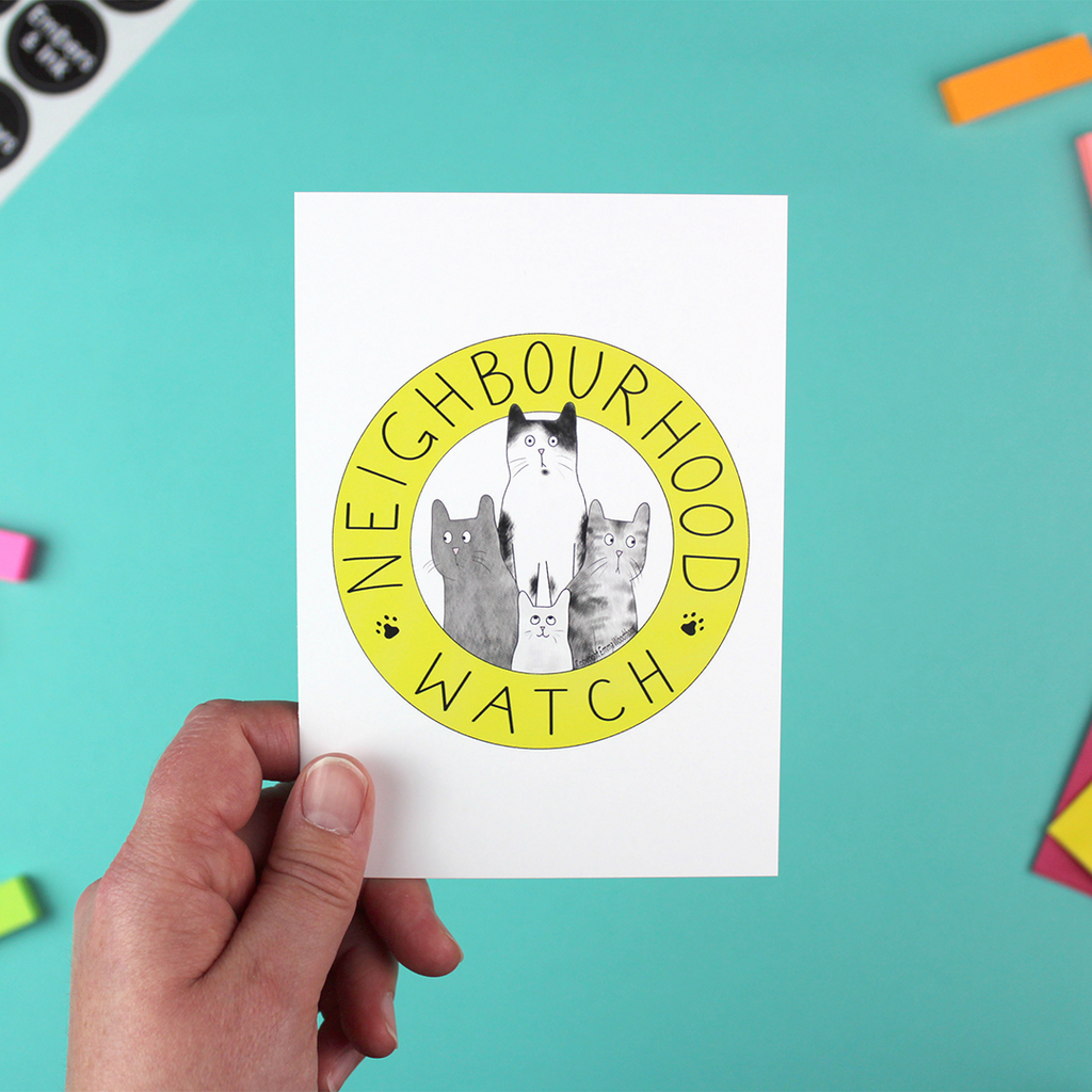 A hand holds up a portrait orientated postcard with the yellow and black neighbourhood watch logo on it, but instead populated by cats!