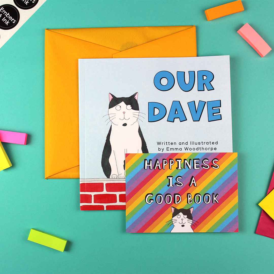 Our Dave is shown next to a wrapped book (in yellow tissue) and a rainbo coloured postcard that has Dave the Cat's face on it, under the words 'happiness is a good book'