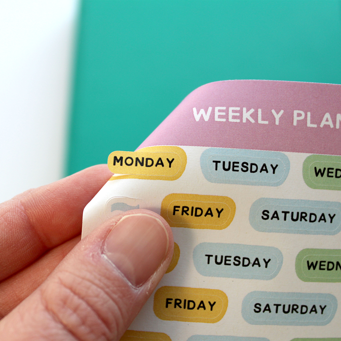 A hand is shown easily peeling the Monday sticker off the sticker sheet.