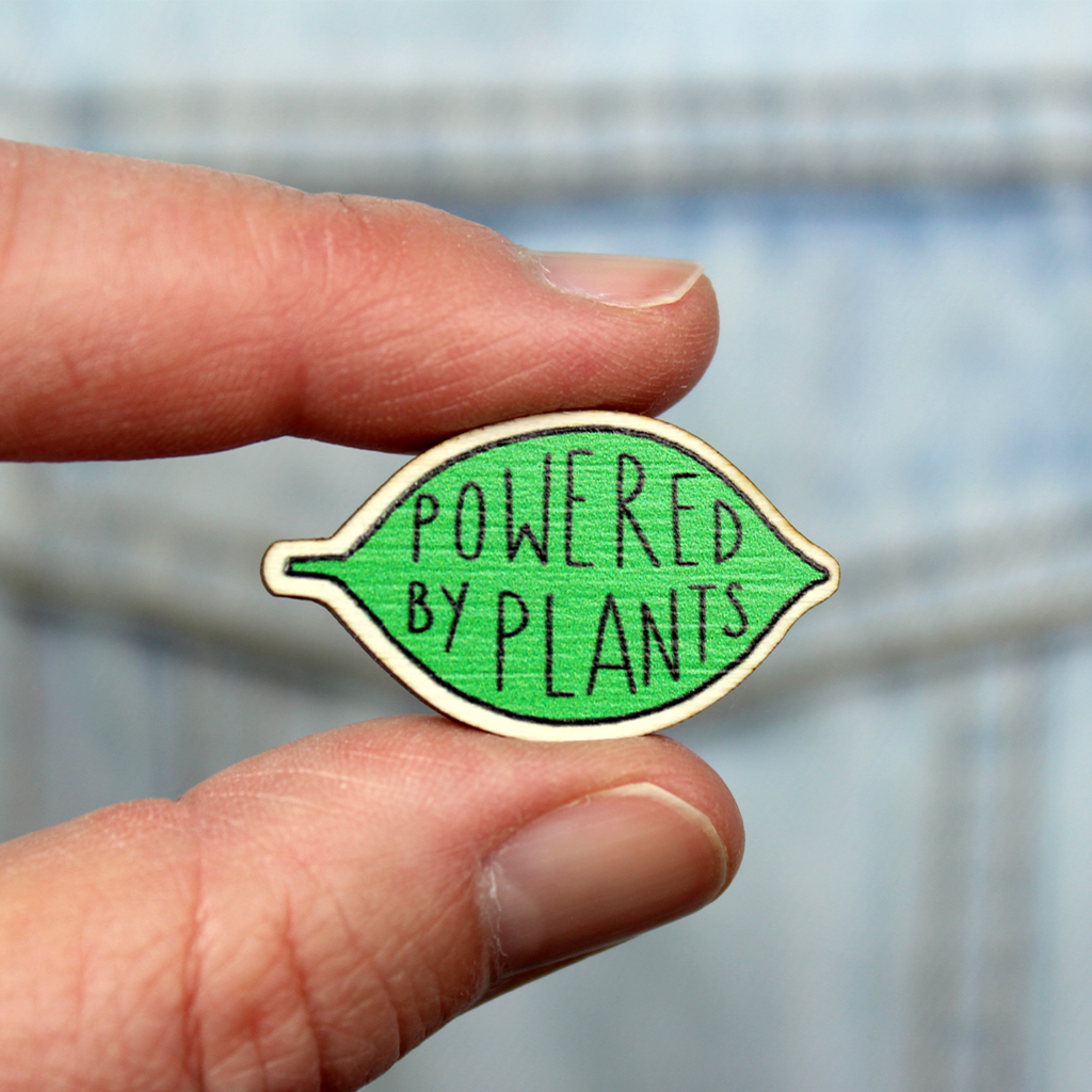 A wooden pin badge in the shape of a green leaf contains the words Powered by Plants. The badge is held between finger and thumb in front of a denim jacket pocket.
