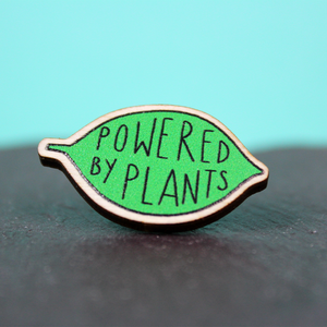 A wooden pin badge in the shape of a green leaf contains the words Powered by Plants. it is resting on a piece of slate.