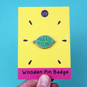 A wooden pin badge in the shape of a green leaf contains the words Powered by Plants. The badge is on a tellow and pink backing board with the words 'wooden pin badge' along the bottom.