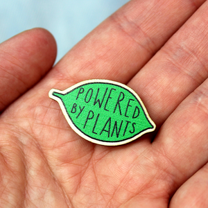 A wooden pin badge in the shape of a green leaf contains the words Powered by Plants.  It is photographed in an open hand.