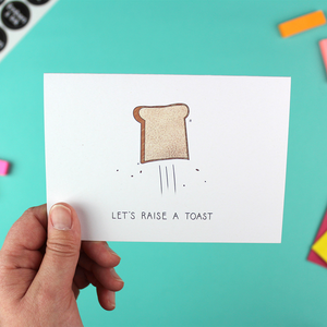 A hand holds a landscape orientated greetings card with an illustration of a piece of toast flying through the air. Underneath are the words Let's raise a toast'