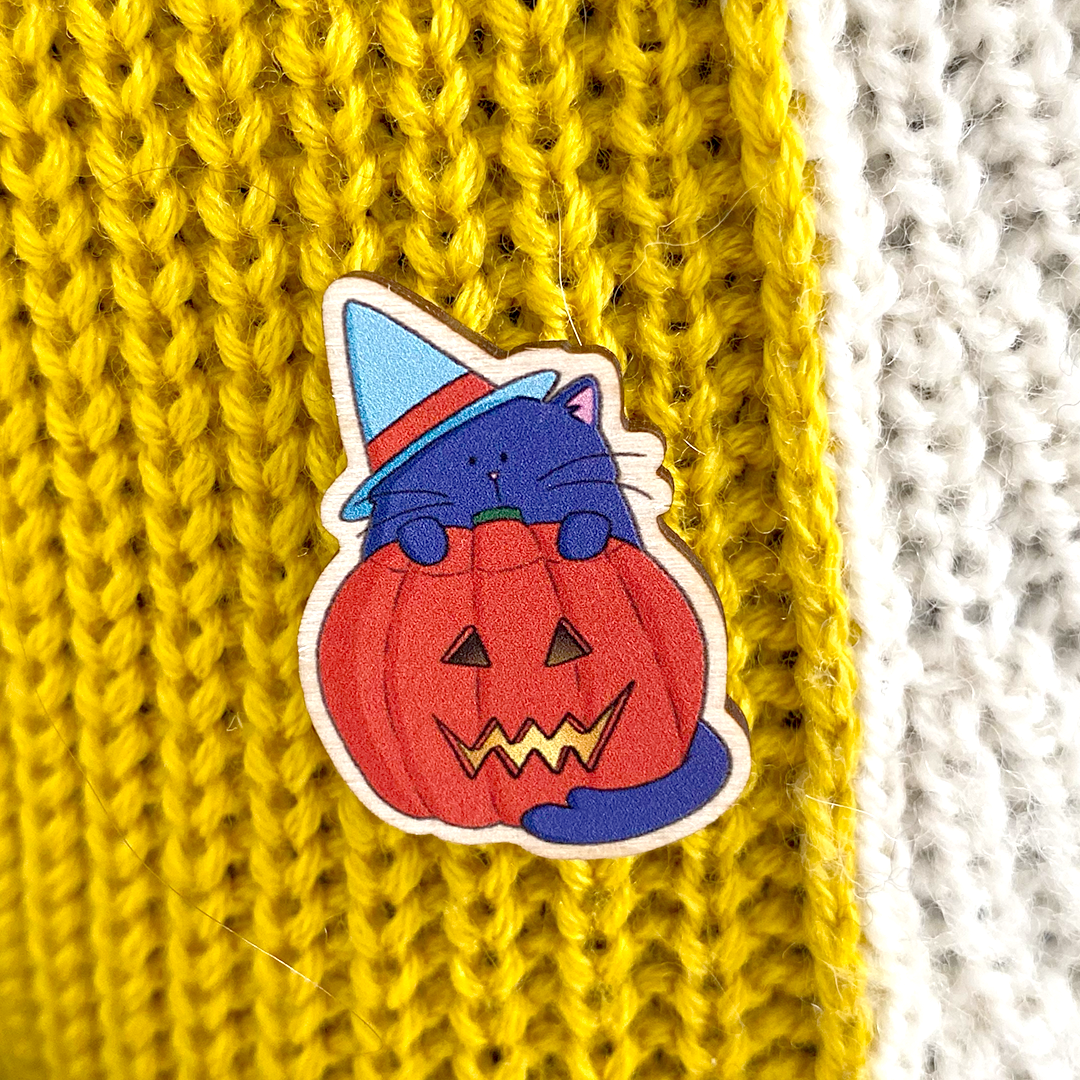 a wooden pin badge featuring an illustrated purple cat wearing a blue witches hat and standing behind a lit spooky halloween pumpkin is shown on a wooly jumper