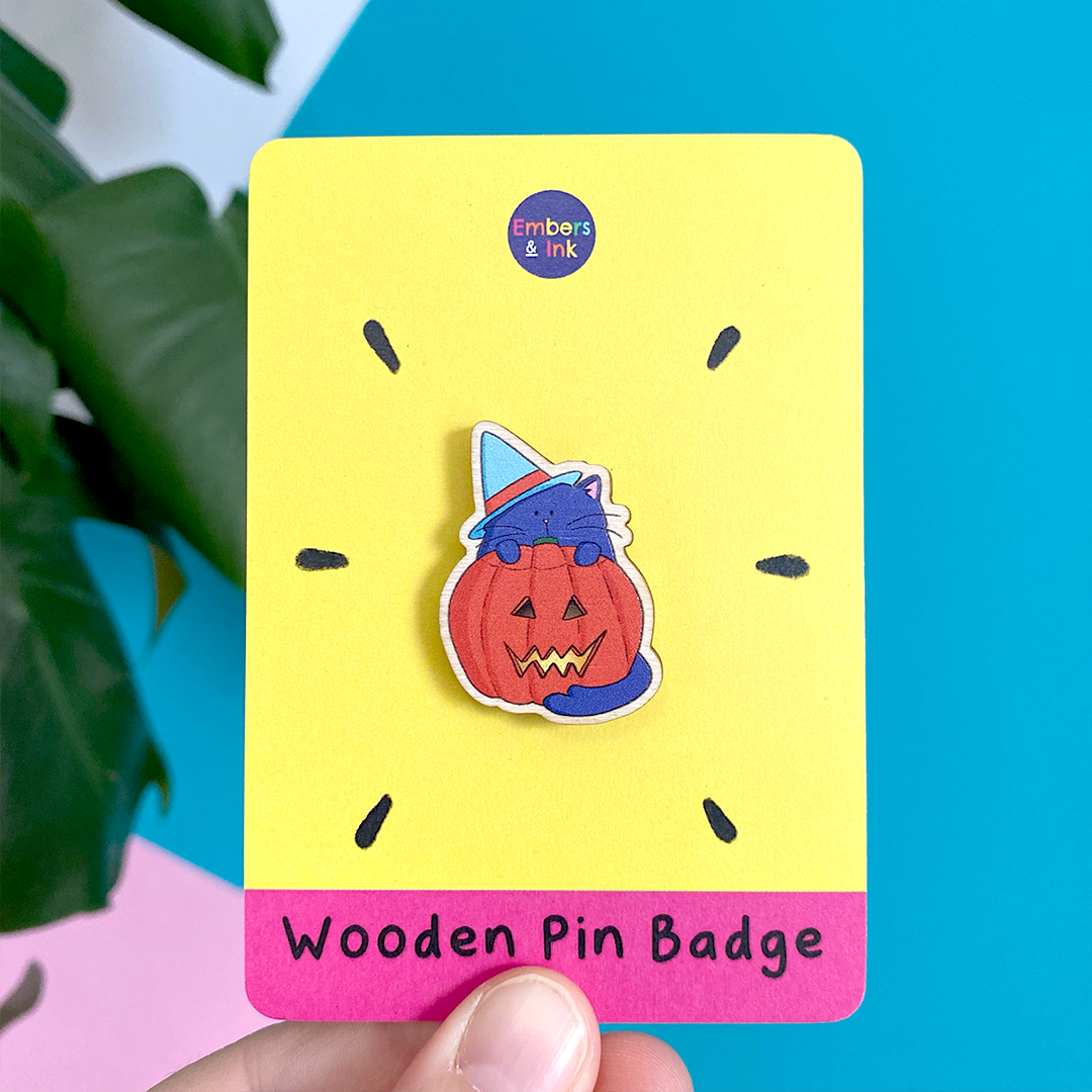a wooden pin badge featuring an illustrated purple cat wearing a blue witches hat and standing behind a lit spooky halloween pumpkin is shown on a pink and yelolow cardboard backer with the words 'wooden pin badge' at the bottom. It is held in front of a brightly coloured wall and a houseplant.