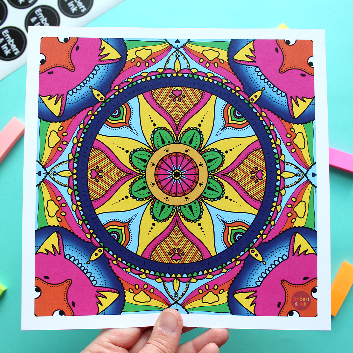 A colourful square art print is shown against a turquise background. The print is a colourful mandala repeating pattern featuring feline things, like cat noses, whiskers, paws, ears and more all hidden in the patternn