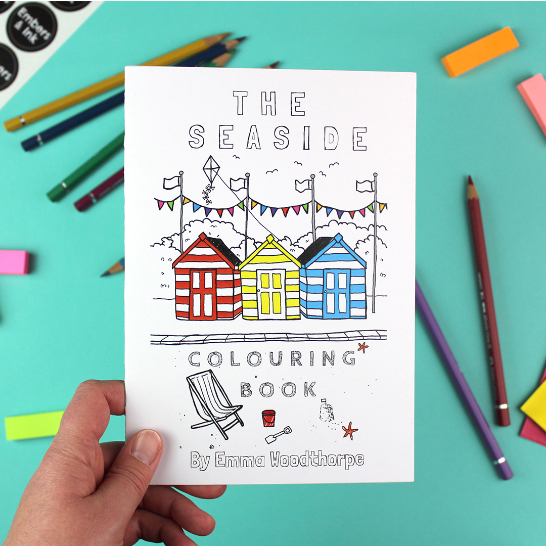 A hand holds the Seaside colouring book. The front cover has a half-coloured illustration of a beach scene with deckchair, sand-castles, beach huts, bunting and flags.