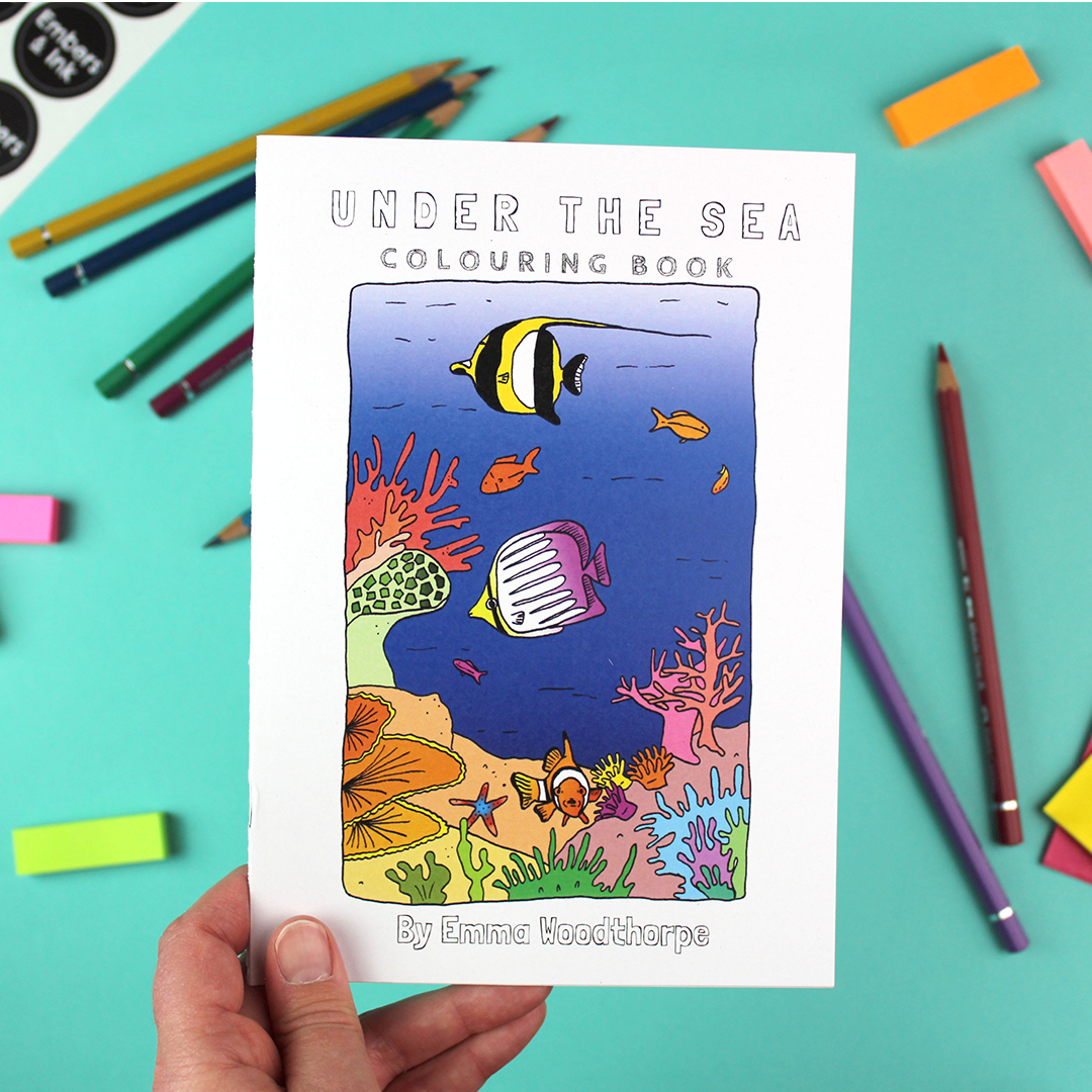 A hand holds the Under the Sea colouring book. the front cover has an illustration of a colourful coral-reef scene.
