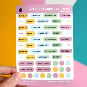 A hand holds up a sheet of Weekly Planner stickers in front of a colourful wall. Each sheet has 4 week day stickers, 4 weekly goes stickers, 8 mini flag decorative stickers and 14 decorative dot stickers.