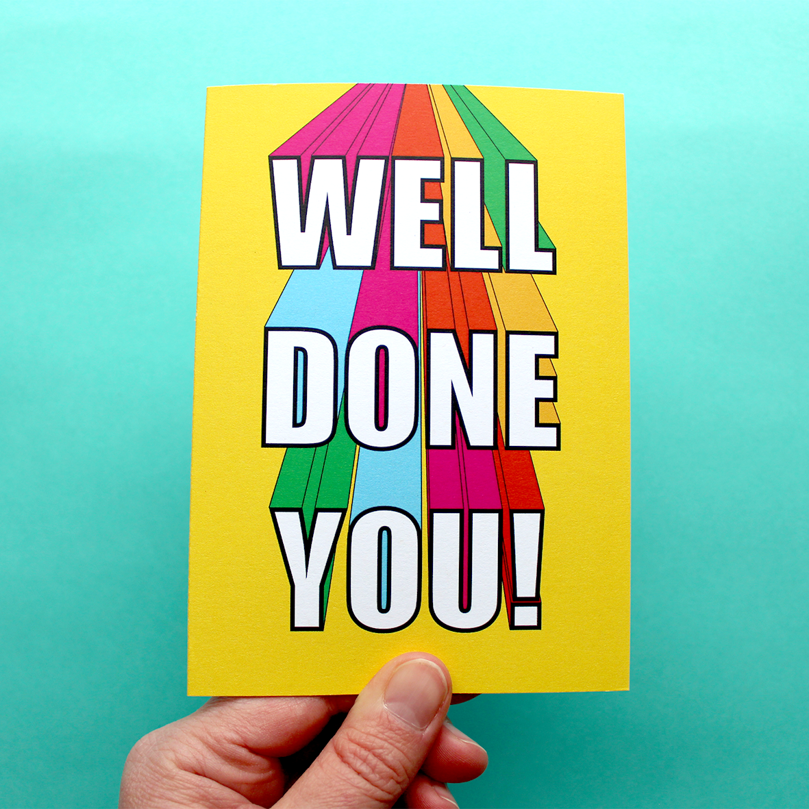 A hand holds a colourful card with the words 'well done you!' on it. The words are in white bubble writing, which is 3D. The words look like they are coming at you from the top of the yellow card. The sides of the 3D writing is rainbow colours.
