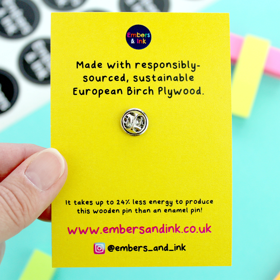 The rear of the cardboard backer is shown. it is yellow with black writing that reads:' made with responsibly-sourced sustainable european birtch plywood. It takes up to 24% less energy to produce this wooden pin than an enamel pin! www dot embers and ink dot co dot uk. instagran @ embers underscore and underscore ink.