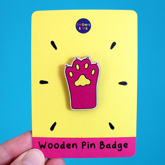 A hand holds a pin paw wooden pin badge that is displayed on a pink and yellow cardboard backer. At. the bottom, on the cardboard backer, are the words Wooden Pin Badge. the Pink cat paw badge has yellow pads and white claws. It is cute!