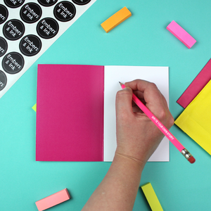 A hand is holding a pink pencil with the embossed white words 'write your own story' on it. there is a notebook, and the hand is poised to start writing.