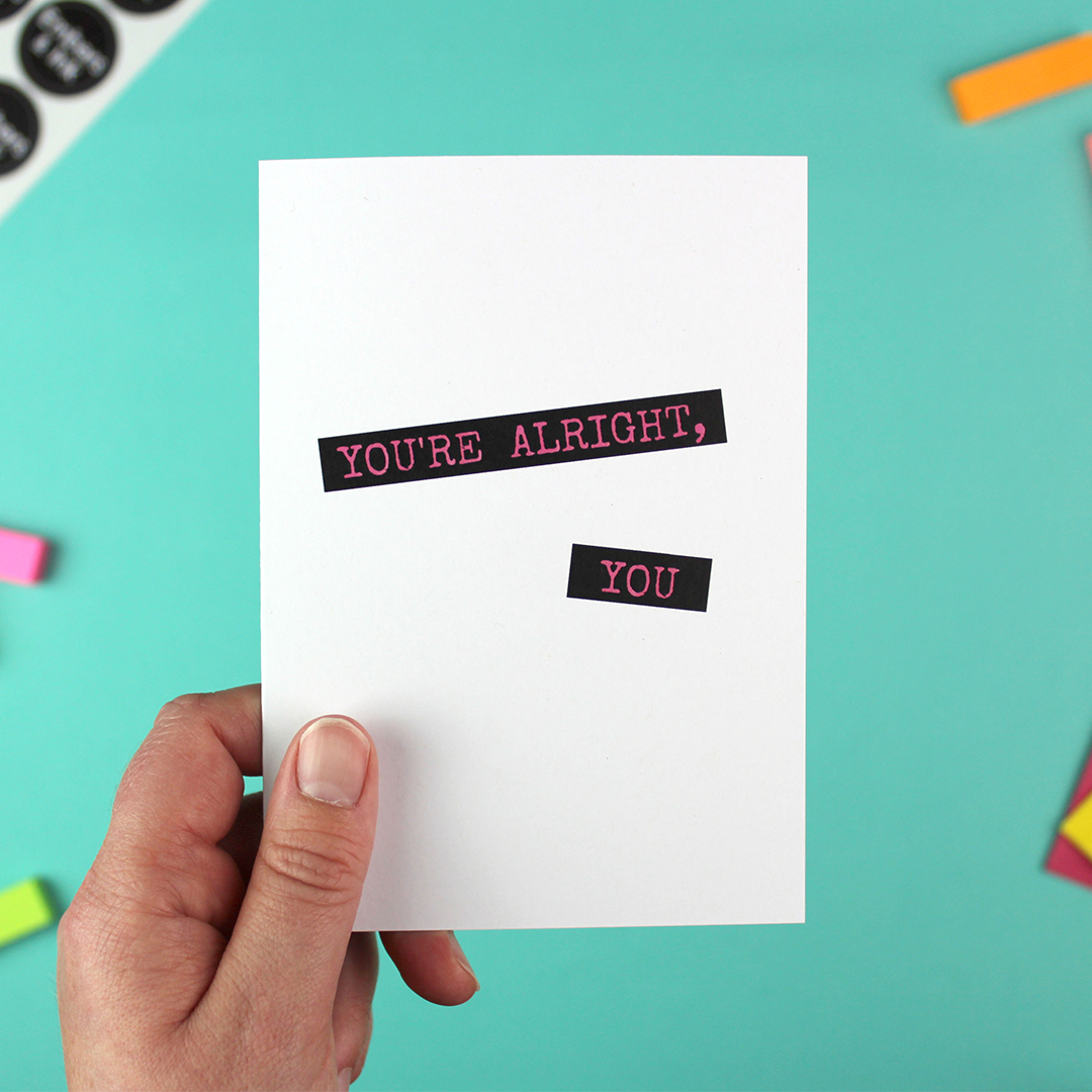 A hand holds a white card with a text effect like a dymo-label writer. The words say 'You're alright, you' with pink text on a black box background.